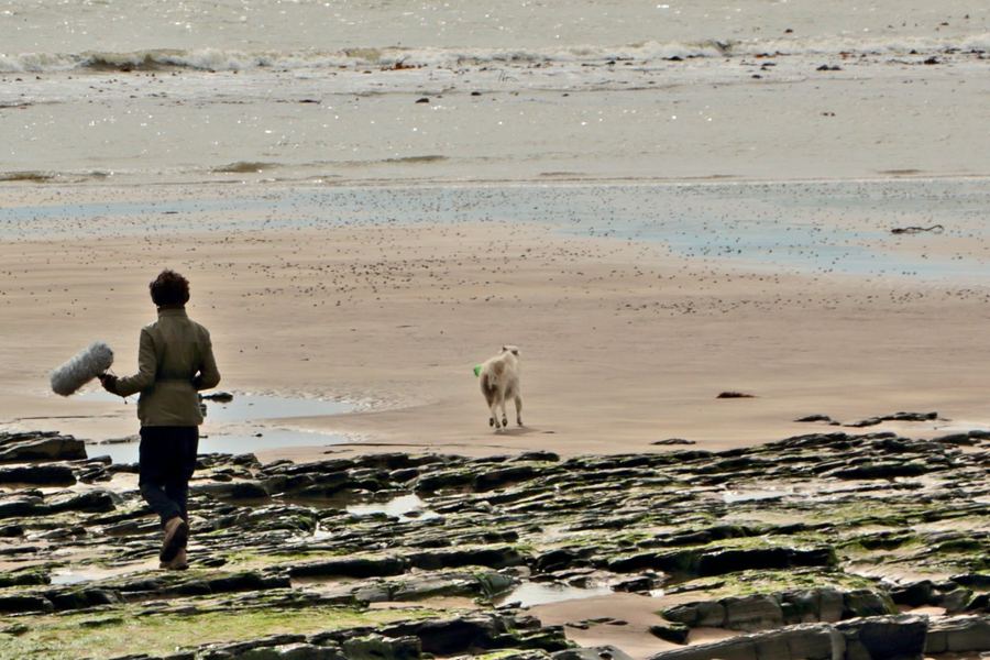 Jason walking across rocks on a beach with a boom microphone, a white dog running ahead towards the sea